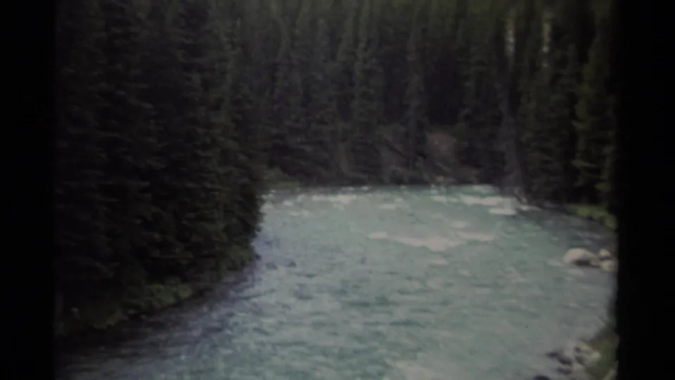 1979 British Columbia 8mm frame with a fast river running past tall conifers, small rocks, and a dark vertical film edge.