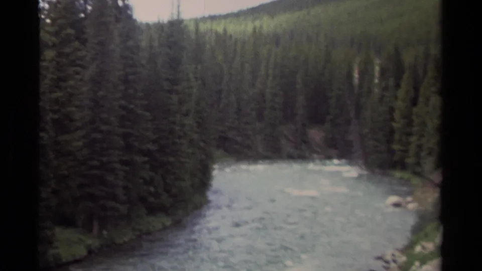 1979 British Columbia 8mm frame showing an evergreen-lined river below a wooded mountain slope, with soft exposure and visible film-frame borders.