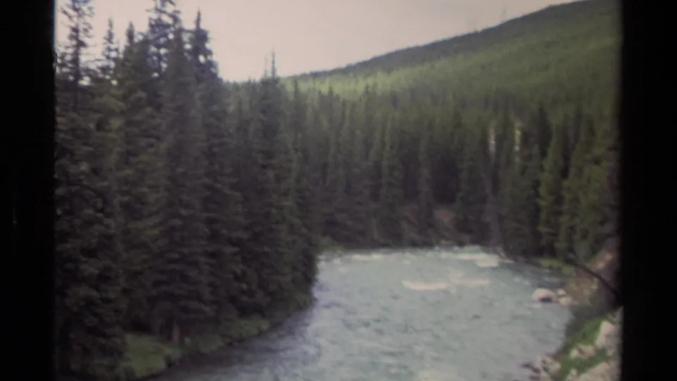 1979 British Columbia 8mm frame looking over a pale mountain river bordered by dense conifers and a sloping forested hillside.