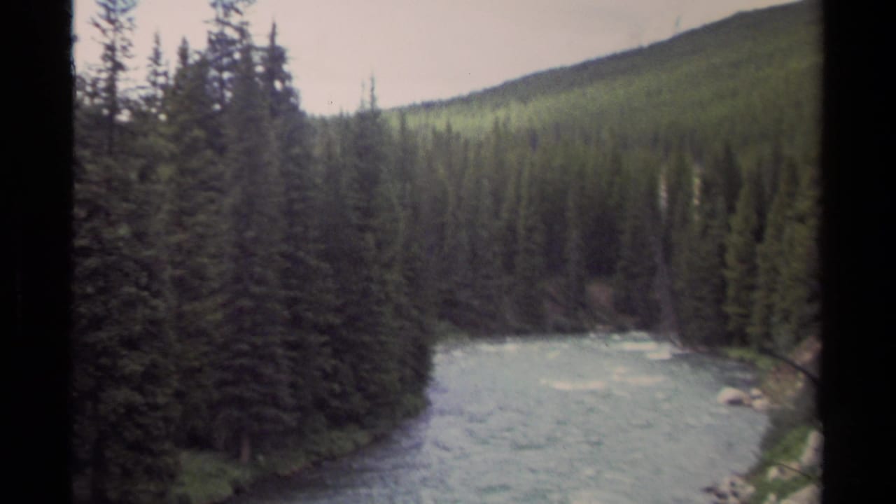 1979 8mm view in British Columbia showing a fast river bordered by dark conifers and rising forested terrain.