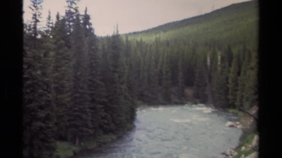 1979 8mm view in British Columbia showing a fast river bordered by dark conifers and rising forested terrain.