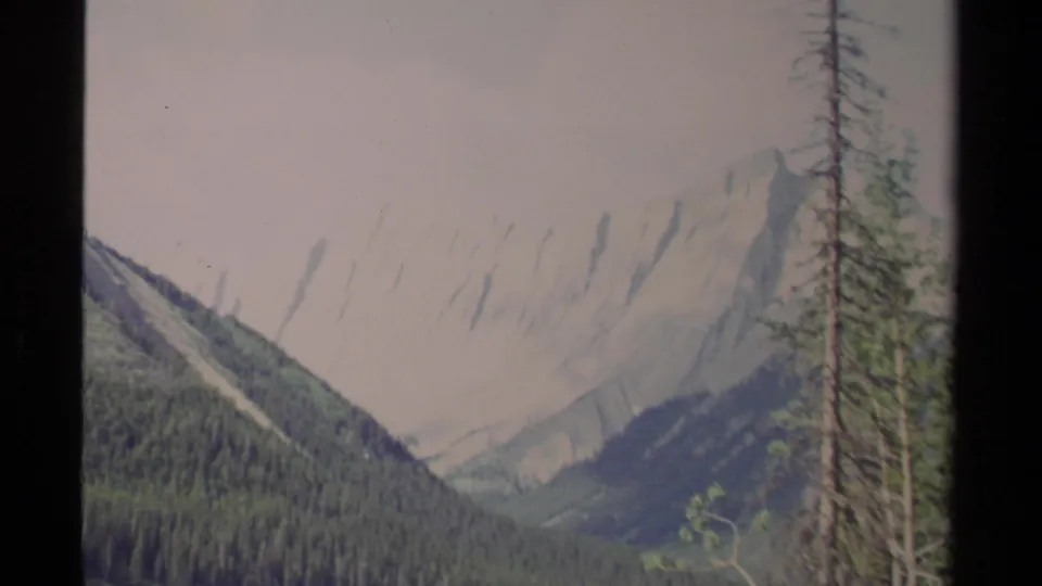 British Columbia mountain valley on 8mm film, with conifer-covered slopes leading toward pale rocky peaks.