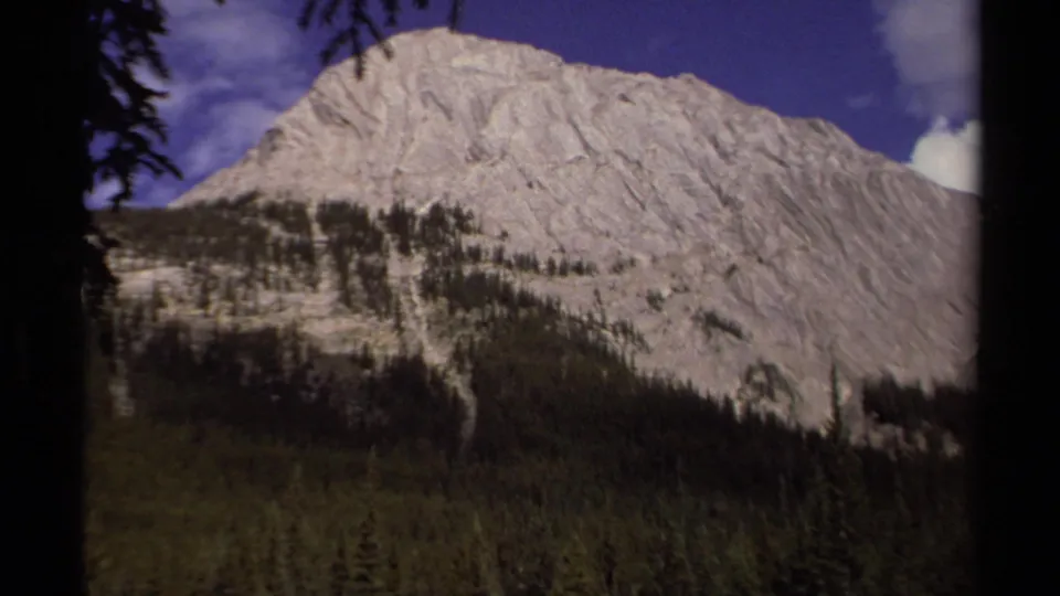 Vintage 8mm view of a rocky mountain ridge and conifer forest in British Columbia, with strong color shift and frame-edge shadow.