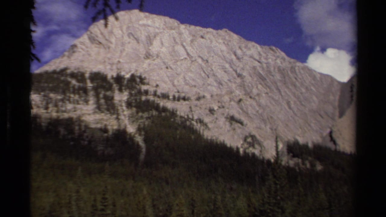 1979 8mm British Columbia mountain view with pale rock slopes, evergreen forest at the base, and scattered clouds in a deep blue sky.