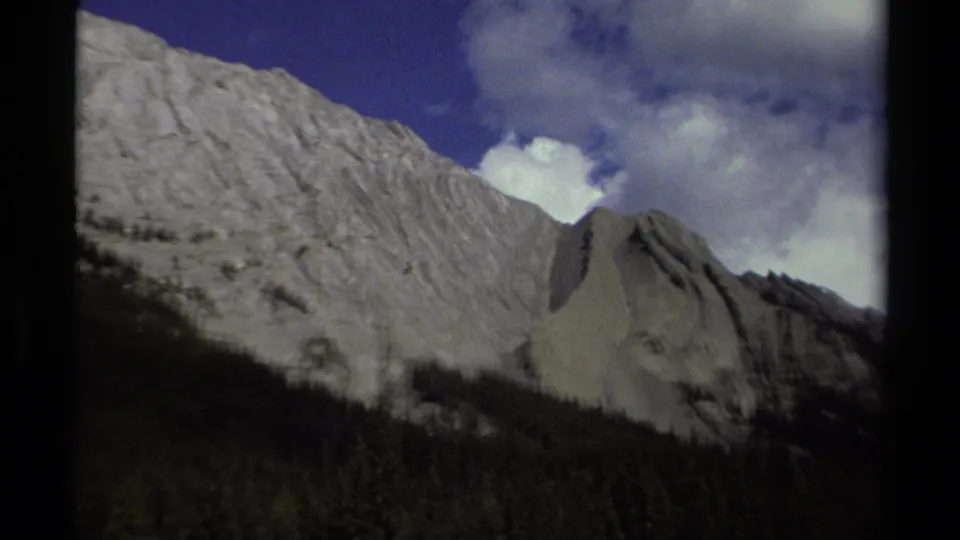 1979 home-movie frame from British Columbia showing rugged mountain slopes, dark forest foreground, and mixed blue sky with clouds.
