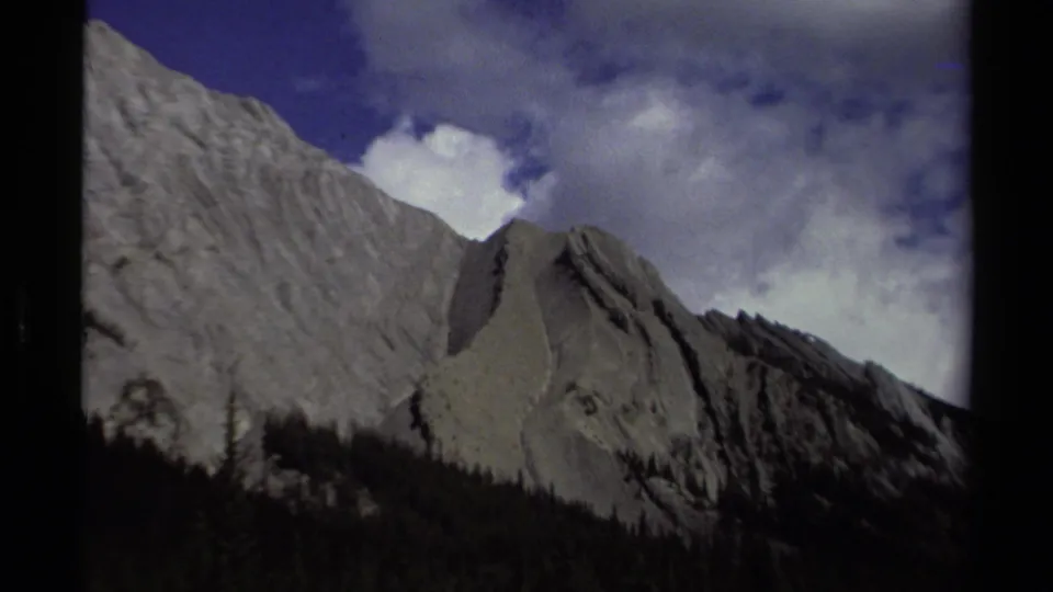 British Columbia 8mm frame showing jagged pale peaks, a dark forest base, blue sky, clouds, and visible film gate edges.