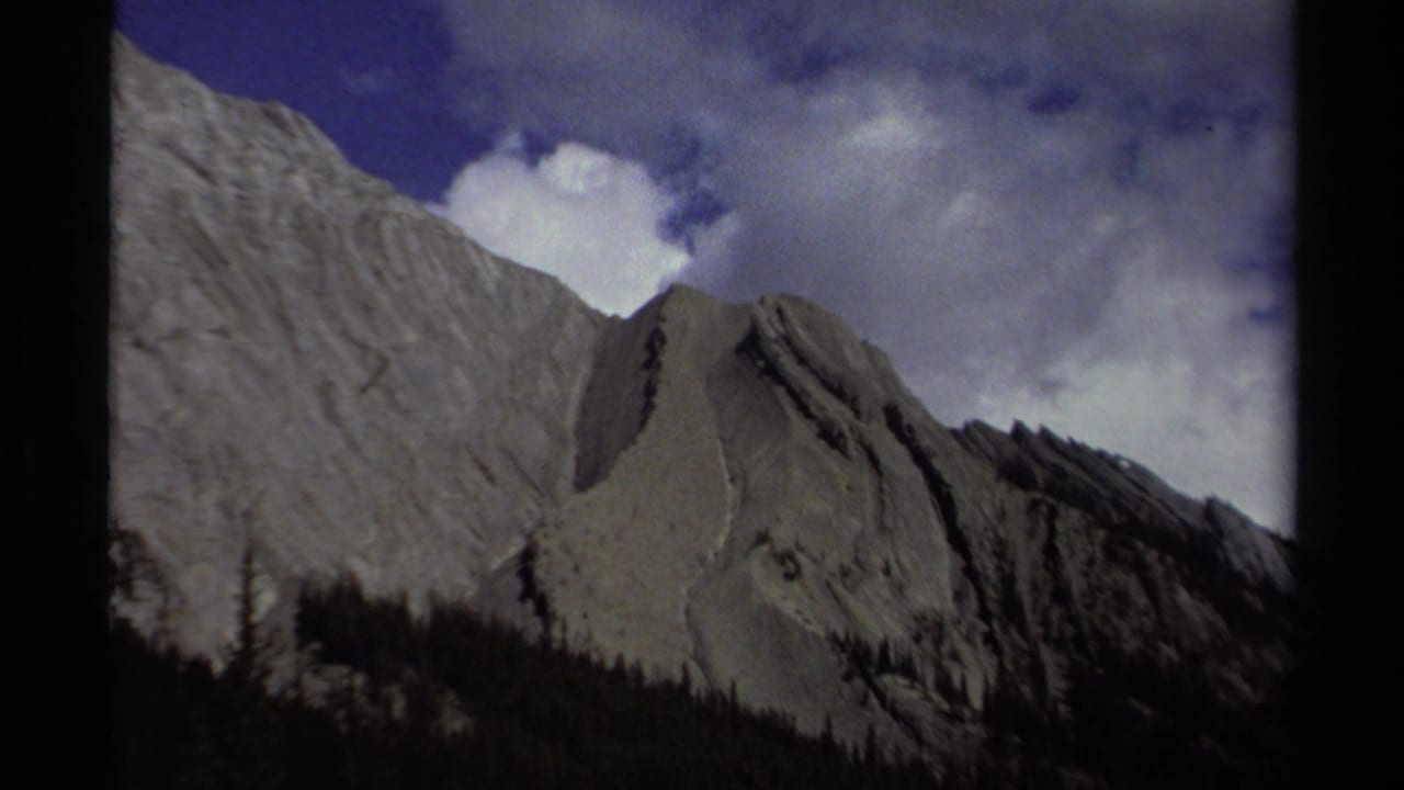 1979 8mm British Columbia mountain ridge with pale rock faces, dark trees, blue sky, and drifting cloud cover.