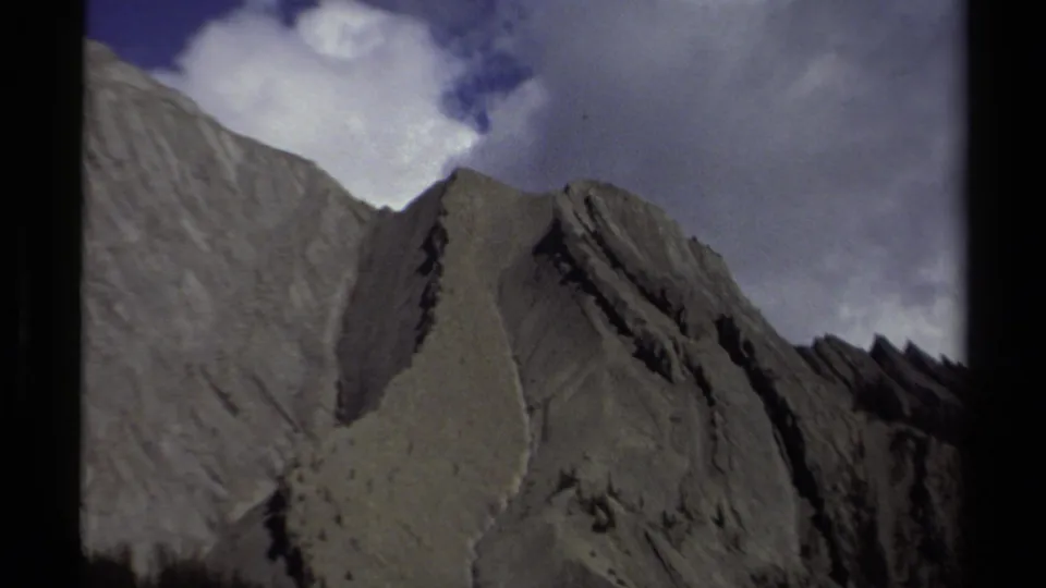 Closer 8mm view of a steep British Columbia ridge, with pale stone, dark fissures, treeline, and cloudy sky.
