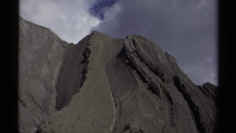 Tight 1979 8mm frame of a British Columbia mountain peak, showing layered rock, shadowed ridges, and cloud cover.