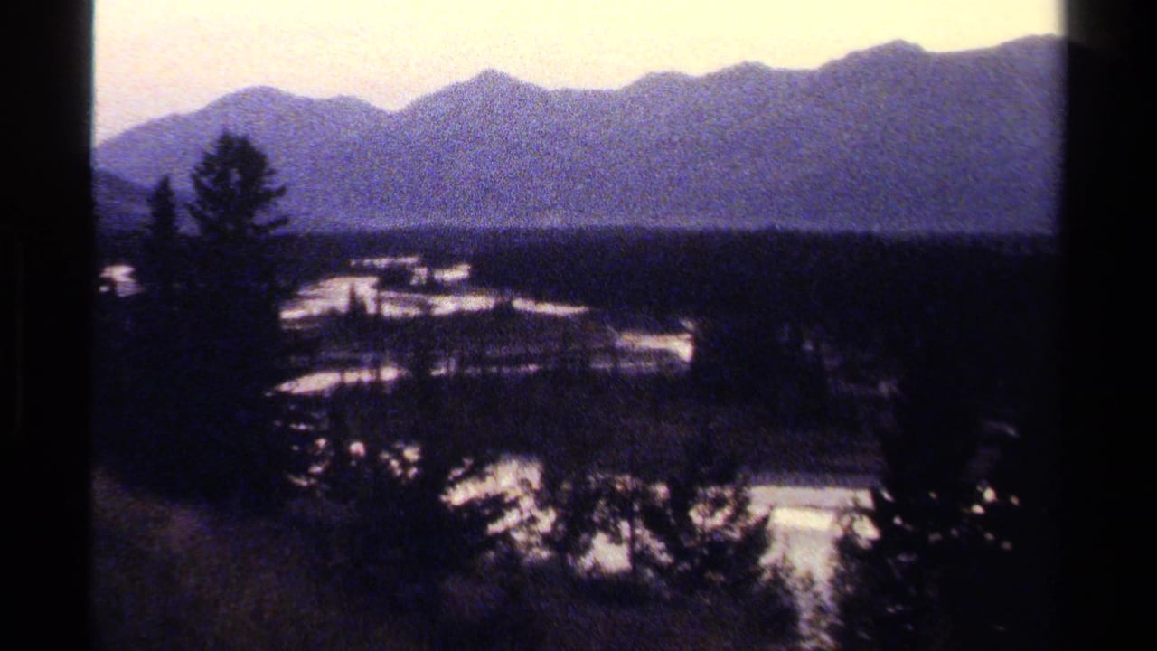1979 8mm view of a British Columbia mountain valley with silhouetted evergreens, pale sky, and snow-bright ground below distant ridges.