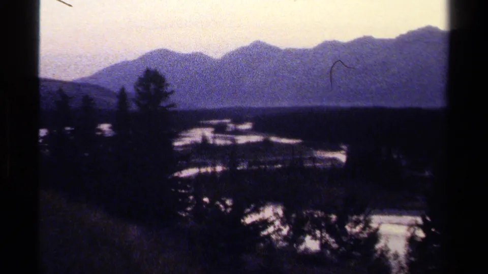 British Columbia mountain landscape on 8mm film, with blue-toned ridges, evergreen silhouettes, and light snow patches across the valley floor.
