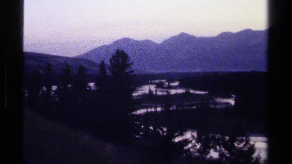 Soft 1979 home-movie frame showing British Columbia ridges beyond dark evergreens and snow-lit open areas in the valley.