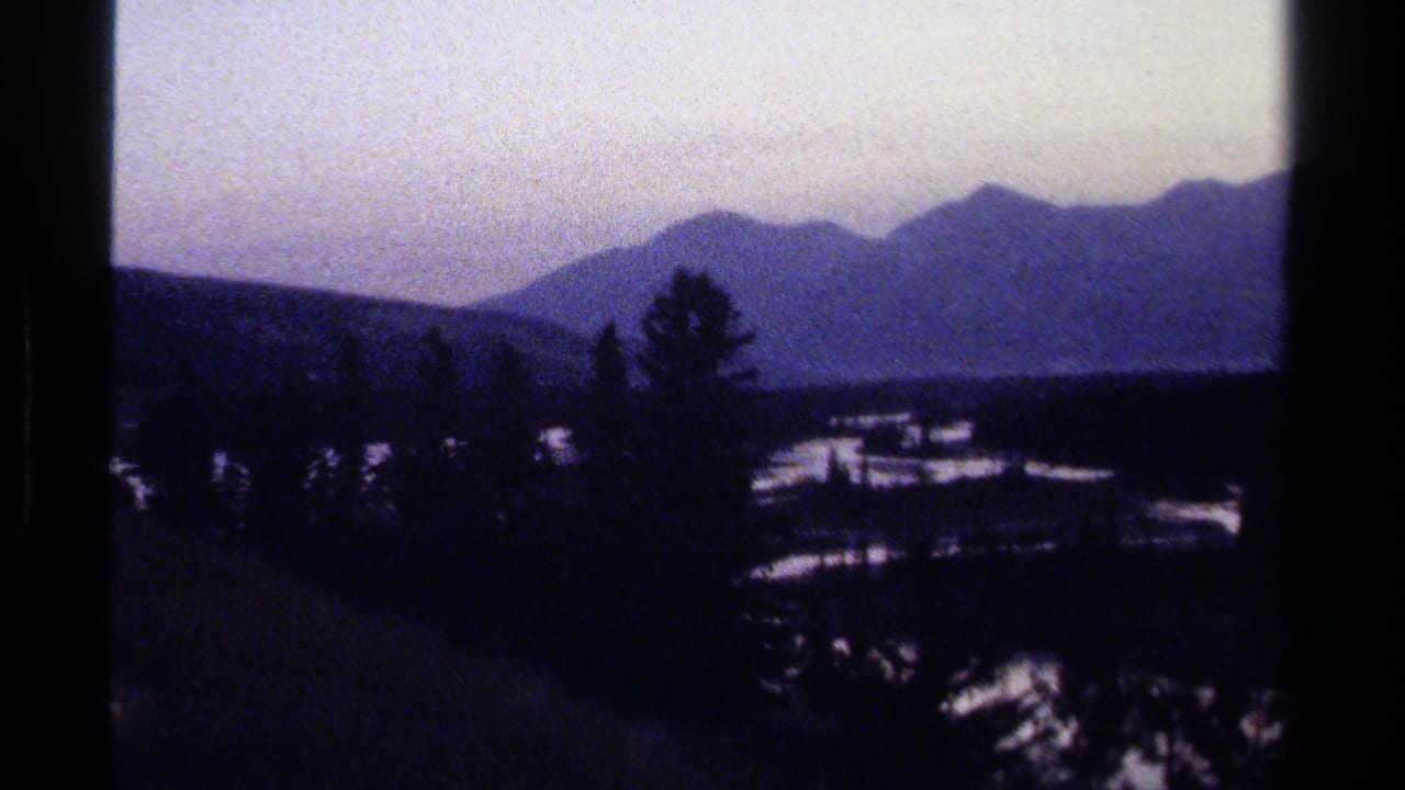 Dark mountain valley with silhouetted trees and pale sky on grainy 8mm film.