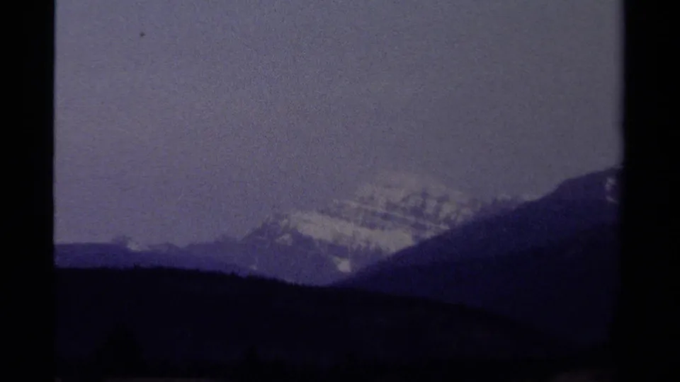 1979 British Columbia 8mm view of a snow-capped mountain rising behind dark forested ridges beneath a muted blue-gray sky.