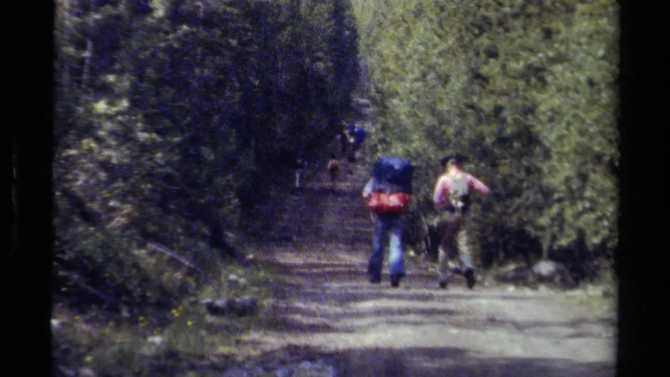 Two backpackers recede along a shaded British Columbia forest road in soft 8mm footage from 1979.