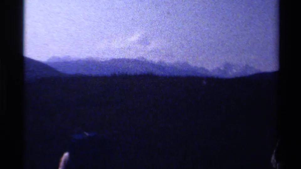 Soft 8mm view of British Columbia mountain ridges and dark forest, with a partial hiker visible near the lower frame edge.
