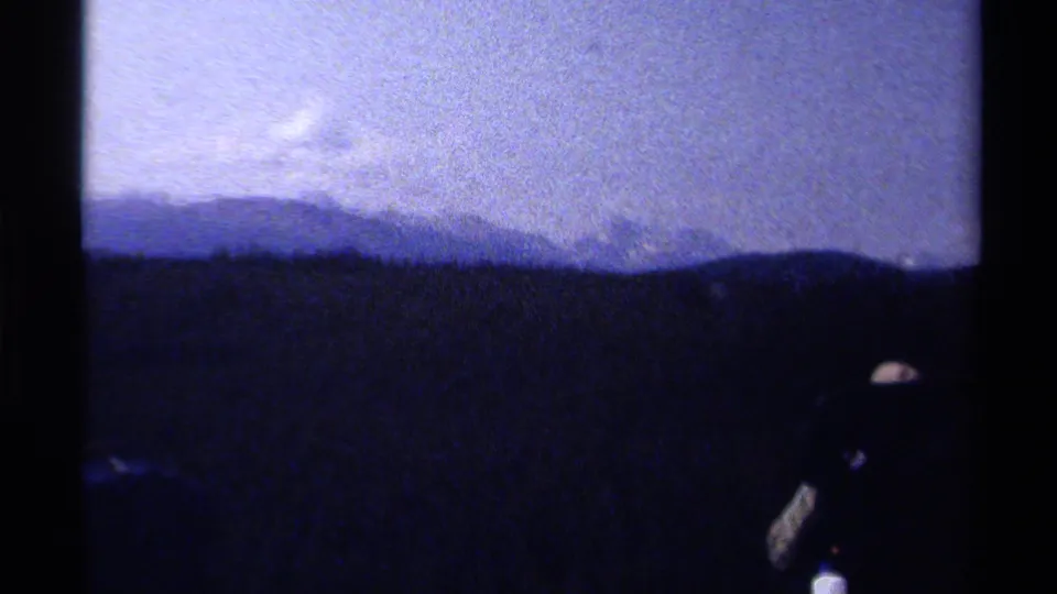 Grainy 8mm frame of a mountain ridge and pale sky, with a backpacked hiker silhouette near the lower right.