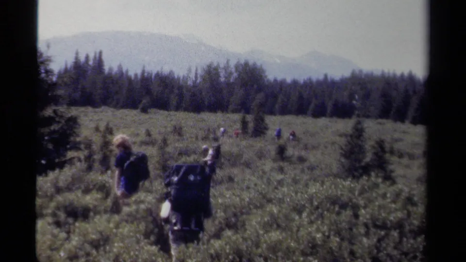 Final 1979 British Columbia frame of hikers with heavy packs crossing brushland, framed by conifer woods and hazy mountains.
