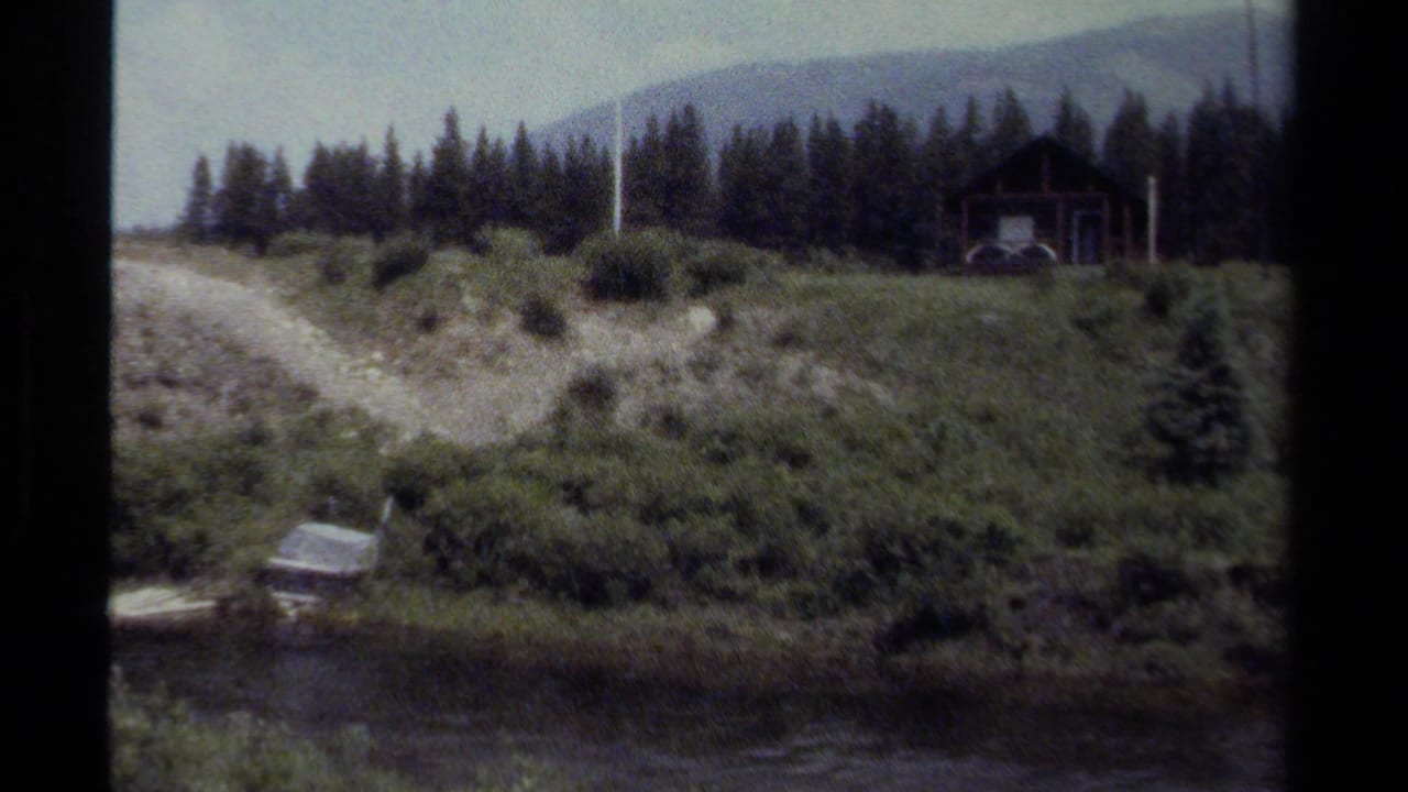 1979 British Columbia home-movie view of a hillside cabin area with a dirt path, Canadian flag, forest line, and mountains beyond.