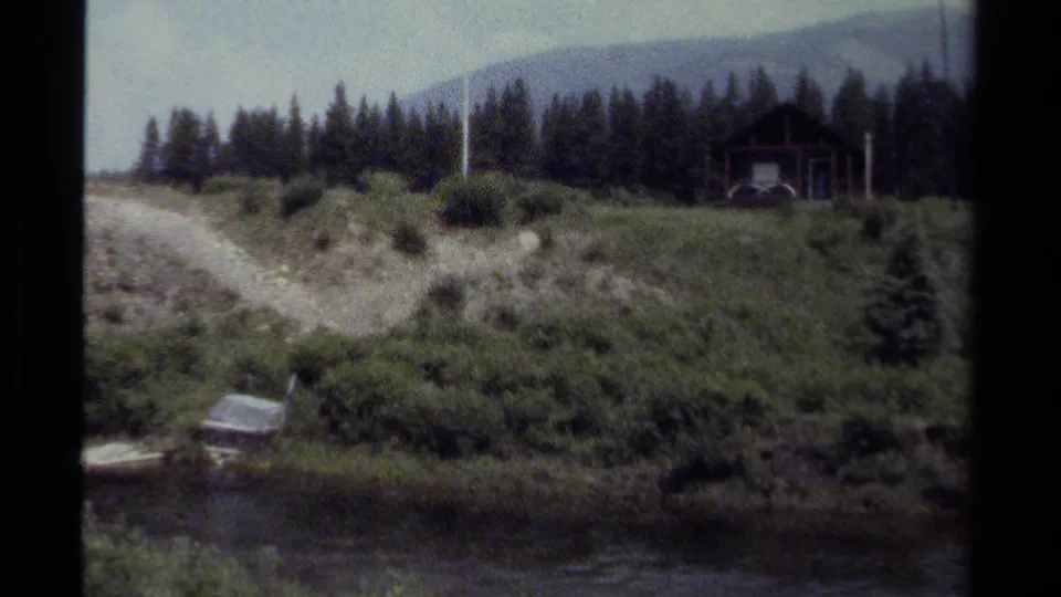 1979 British Columbia home-movie view of a hillside cabin area with a dirt path, Canadian flag, forest line, and mountains beyond.