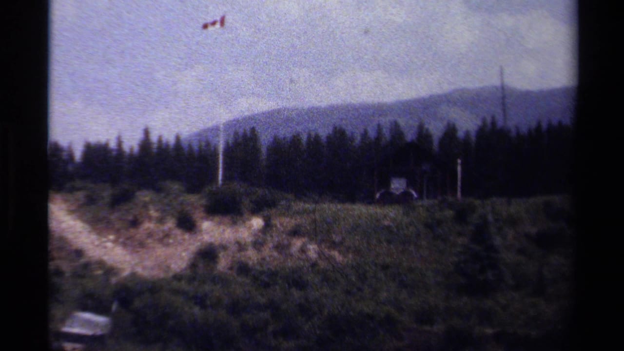 Wide 1979 British Columbia view of a small cabin area on a grassy hillside, with a Canadian flag above evergreens and mountains.