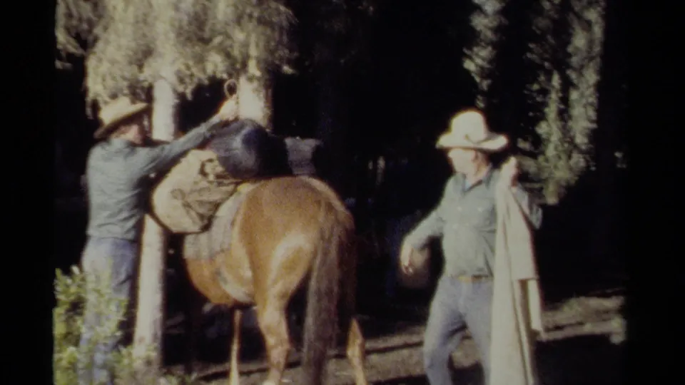A man reaches over the saddled horse as another hat-wearing man stands nearby with gear in the wooded British Columbia setting.