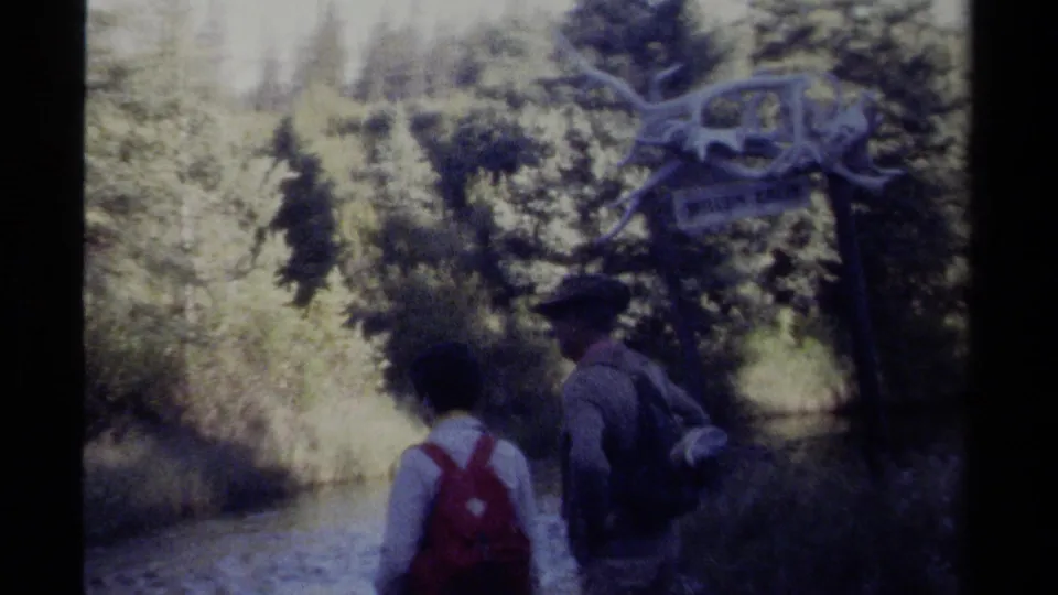 Two backpacked visitors walk along a shallow creek beneath the Willow Creek sign in British Columbia home-movie footage.