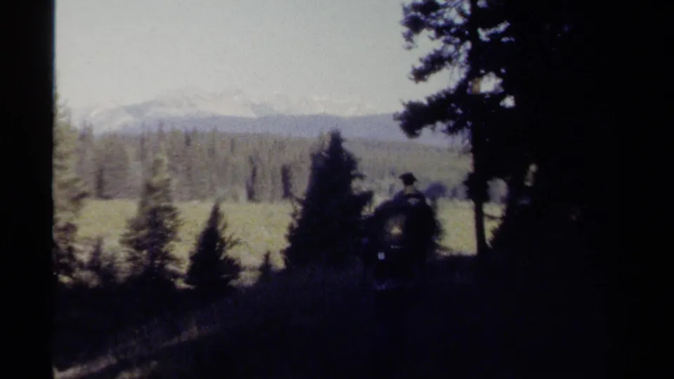 A hiker with a large pack pauses or moves near the trail edge as a forest meadow and distant mountains appear beyond.