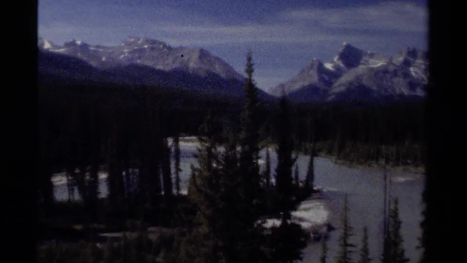 1979 British Columbia landscape frame with snow-capped mountain peaks above a dark evergreen valley, with pale water partly visible near the lower edge.