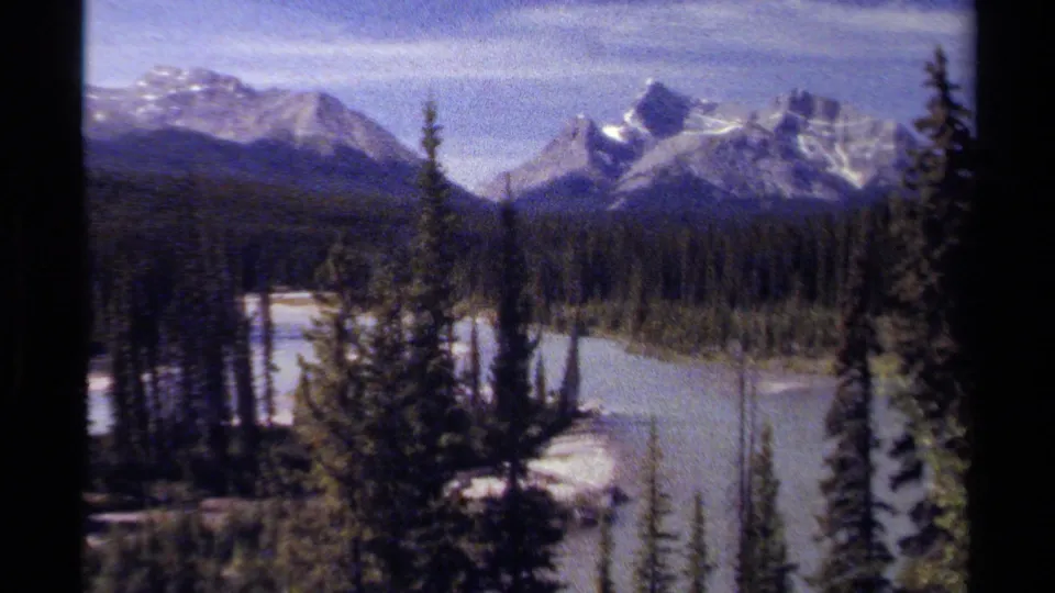 A broader 8mm home-movie view of British Columbia mountains, with snow-bright peaks, evergreen forest, and a pale river channel running through the valley below.