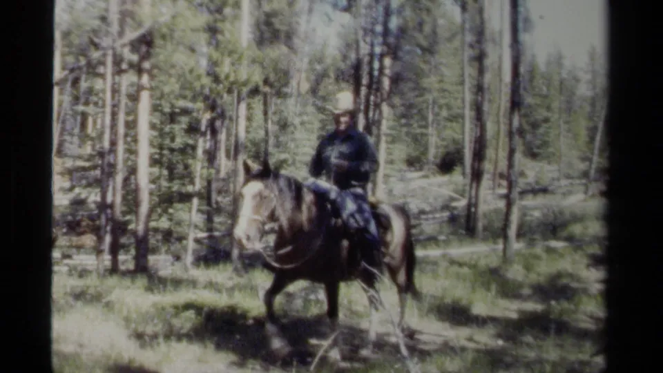 1979 British Columbia frame showing an adult man in a cowboy hat riding a horse across a forested clearing.