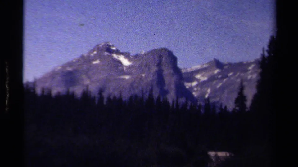 Blue-tinted 8mm frame of a British Columbia mountain peak above dark conifers, with visible soft focus and film border shadows.