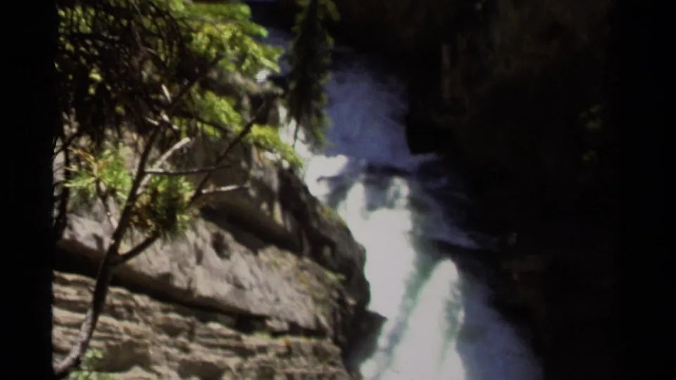 1979 8mm frame of a bright waterfall descending through a shaded British Columbia mountain gorge, partly framed by evergreen branches.