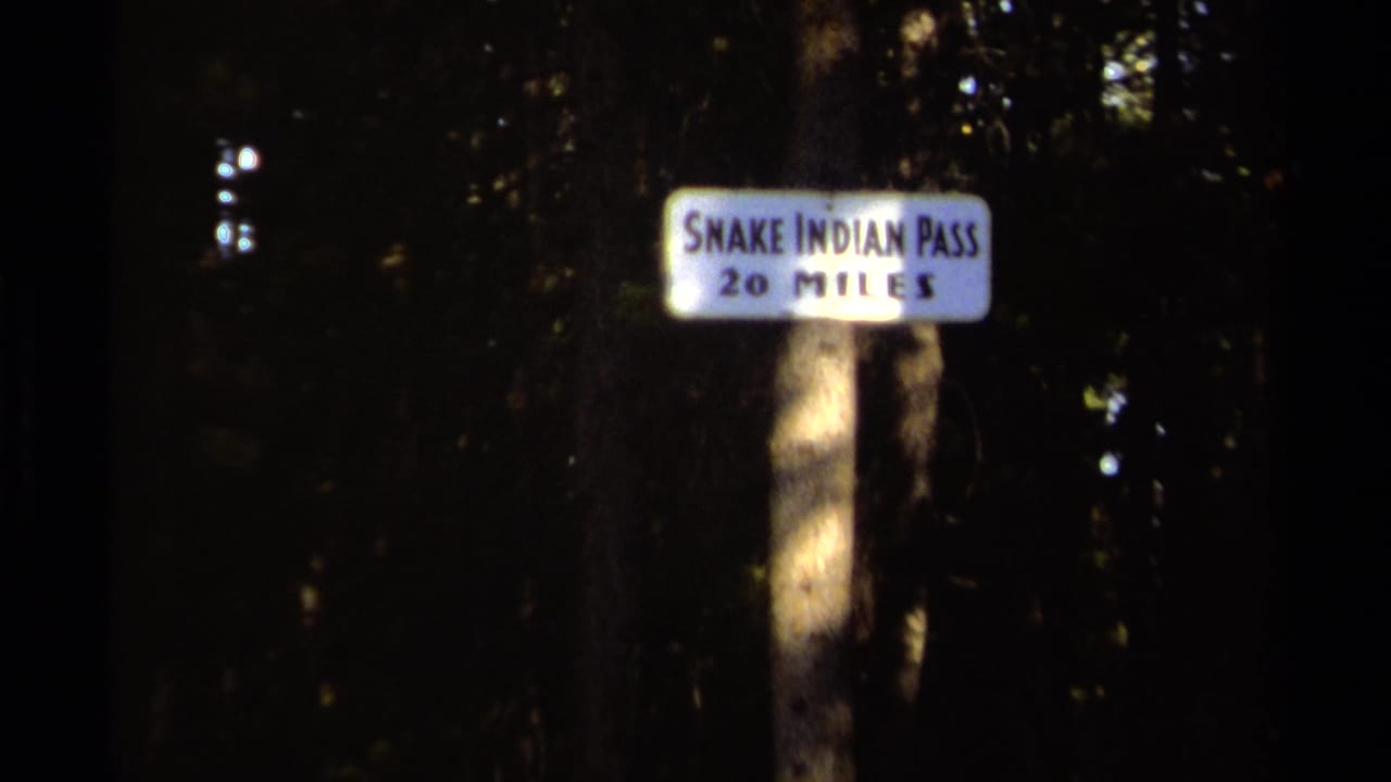 1979 British Columbia trail scene with a white sign reading Snake Indian Pass 20 Miles mounted on a post among dense trees.