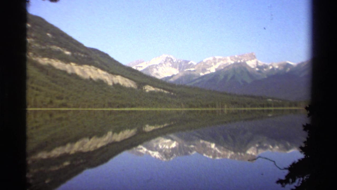 1979 British Columbia 8mm view of a still lake mirroring snow-capped mountains, wooded hillsides, and a clear blue sky.