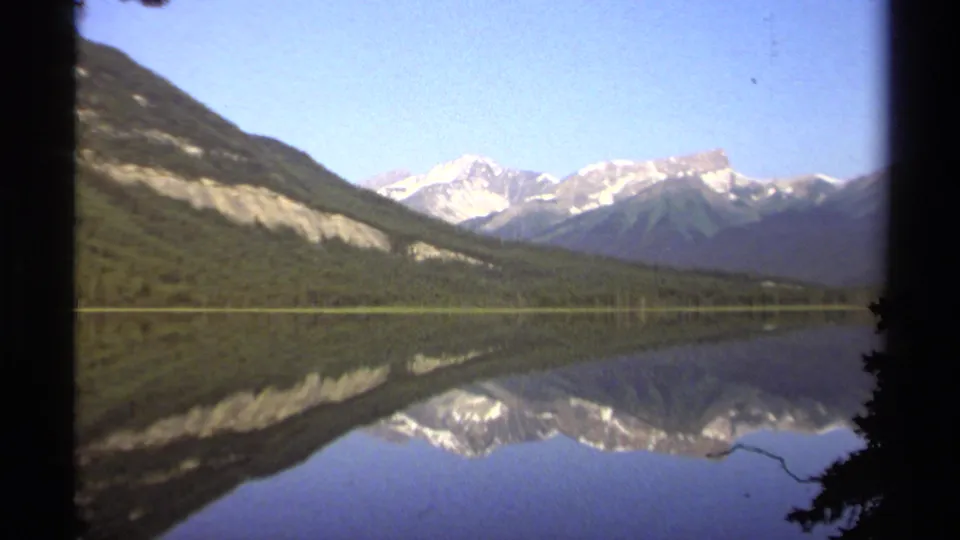 1979 British Columbia 8mm view of a still lake mirroring snow-capped mountains, wooded hillsides, and a clear blue sky.
