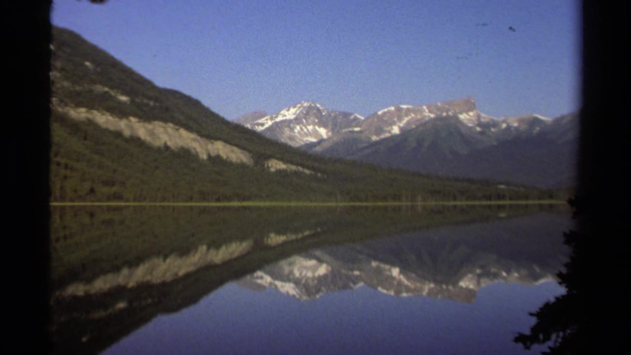 A British Columbia mountain lake reflects snowy peaks and a forested slope in soft 8mm color footage from 1979.