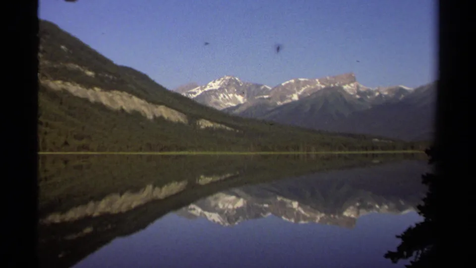 1979 8mm travel footage shows forested slopes, distant snow-capped mountains, and their reflection on a quiet British Columbia lake.
