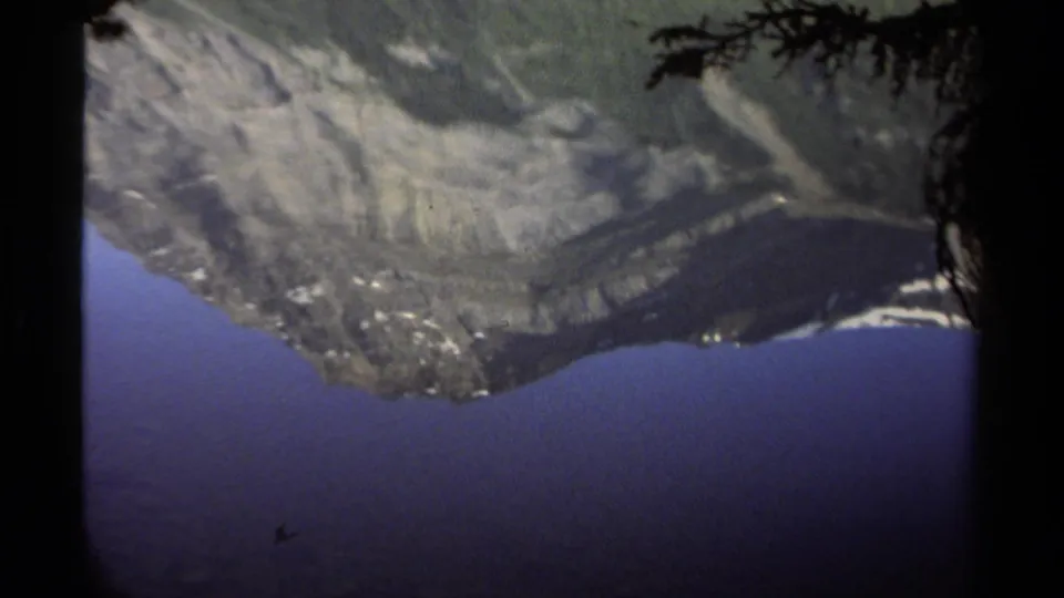 The 1979 British Columbia lake view shifts to an abstract reflection of mountain slopes, blue water, and a dark tree edge in 8mm footage.