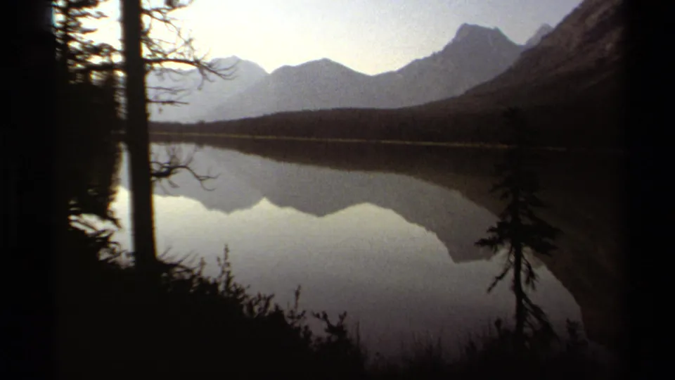 1979 British Columbia home-movie frame of a still mountain lake reflecting peaks, framed by trees and brush.