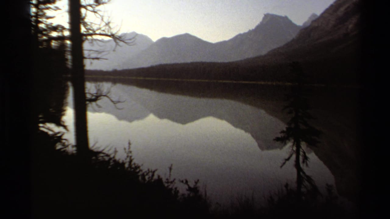 Dark pine silhouettes frame a still lake reflecting distant mountains at low light.