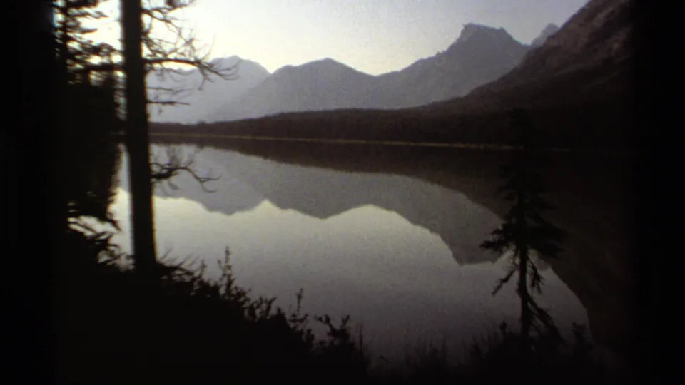 1979 British Columbia 8mm frame with silhouetted lakeshore trees, calm water, and a mountain range reflected under a dim sky.