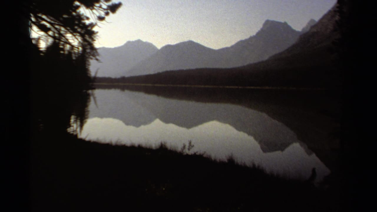Mountain range and lake reflection seen between dark foreground trees.