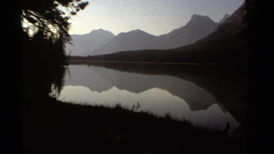1979 British Columbia 8mm frame showing a quiet lake and reflected mountain range partially framed by dark evergreen trees.