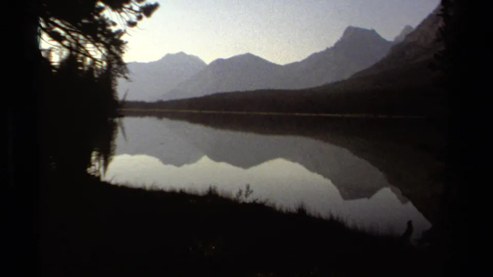 1979 British Columbia 8mm frame with dark lakeshore vegetation, distant mountains, and a broad reflection across still water.