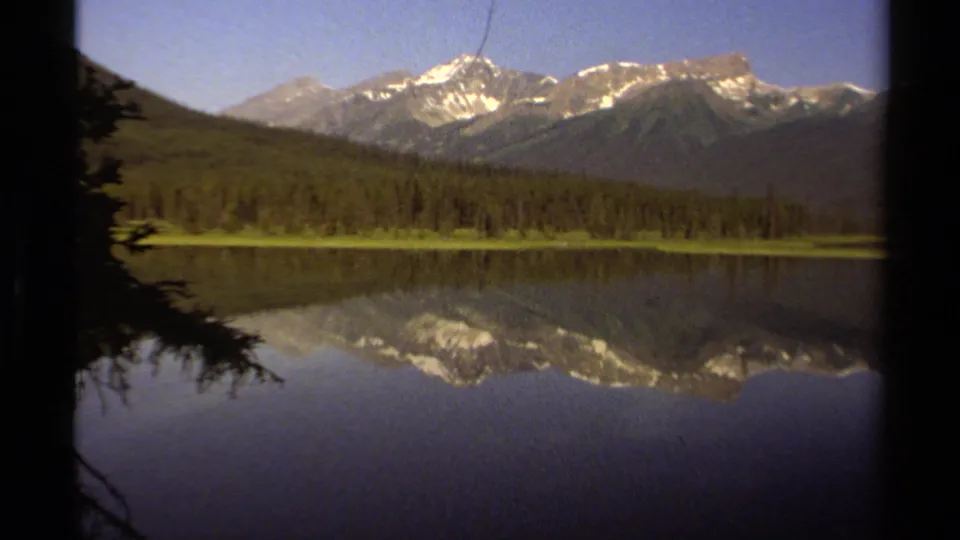 1979 British Columbia 8mm frame showing snow-capped mountains, evergreen forest, and a clear lake reflection in brighter light.