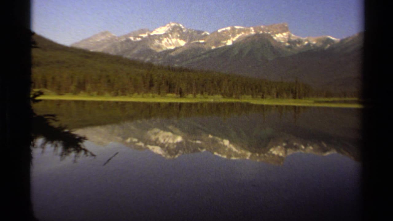1979 8mm view of British Columbia mountains above a forested lake, with snowy peaks reflected in still water.