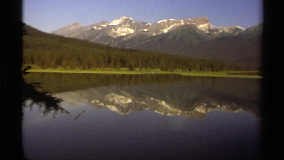 1979 8mm view of British Columbia mountains above a forested lake, with snowy peaks reflected in still water.