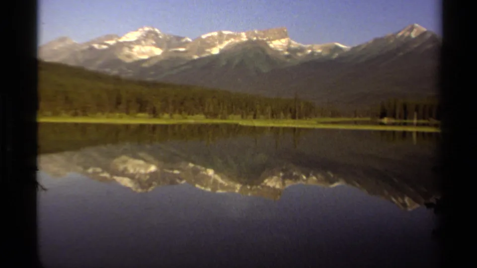 British Columbia mountain landscape on 8mm film, with snowy ridges, a forest line, and mirrored lake surface.