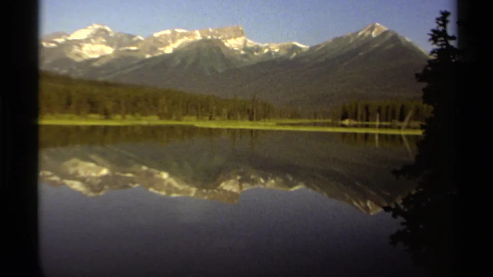 Vintage 8mm British Columbia lake scene with snow-topped peaks, dense evergreens, and a clear reflection across the water.